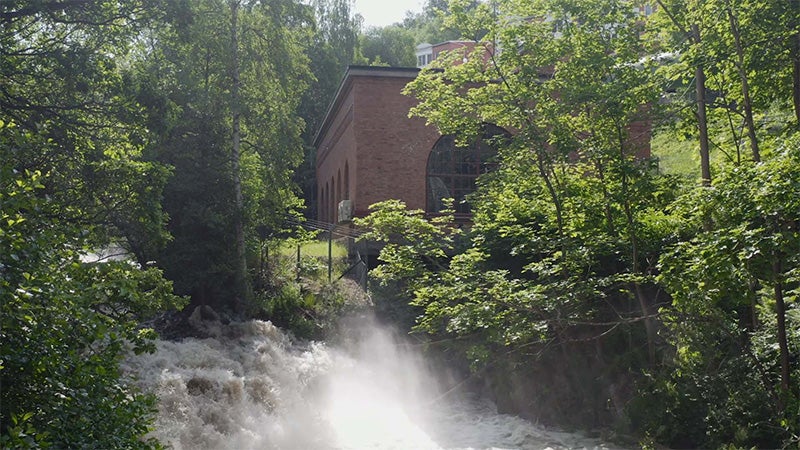 Röd tegelbyggnad från Tabergs kraftstation omgiven av grönskande träd, med vatten som forsar kraftigt nedför berget i förgrunden.