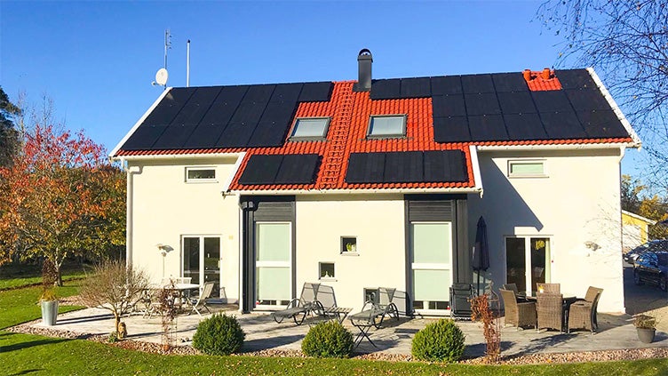 Vit tvåplansvilla med svarta solcellspaneler installerade på taket och en välskött trädgård med utemöbler i höstsolen.