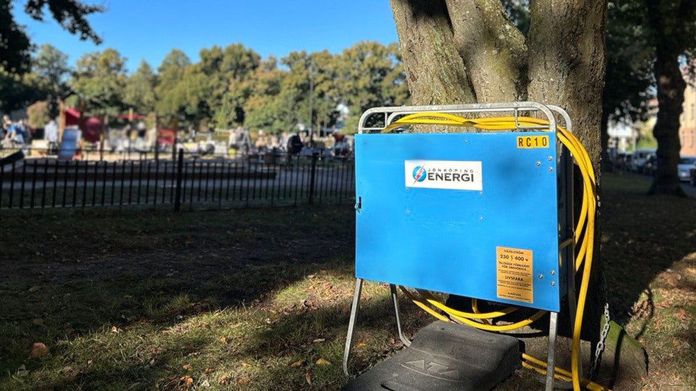 En portabel elcentral från Jönköping Energi med gula elkablar, placerad vid ett träd i en park. Bakgrunden visar en lekplats och träd i dagsljus.