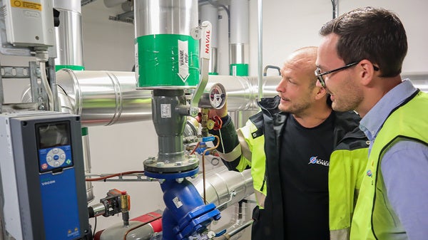 Två män i varselkläder står och inspekterar en mätare på en värmecentral.