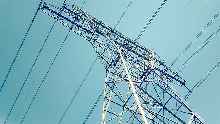 Högt elledningstorn i metall fotograferat underifrån med flera kraftledningar som sträcker sig över himlen, symboliserar elöverföring i elnätet.