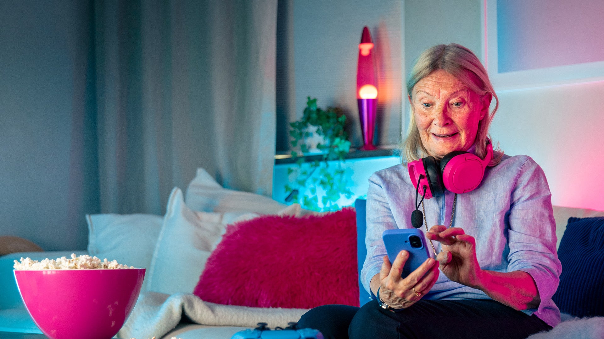 Äldre kvinna med rosa hörlurar runt halsen sitter i en soffa och använder sin smartphone, med en skål popcorn bredvid sig och färgglad belysning i bakgrunden.