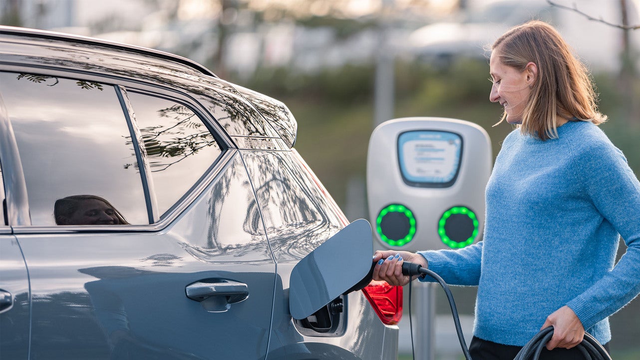 En kvinna i blå tröja ansluter en laddkabel till en grå SUV vid en laddstation. Laddstationens gröna ljus indikerar att den är redo för laddning. Scenen äger rum utomhus med suddig bakgrund av natur och andra fordon.