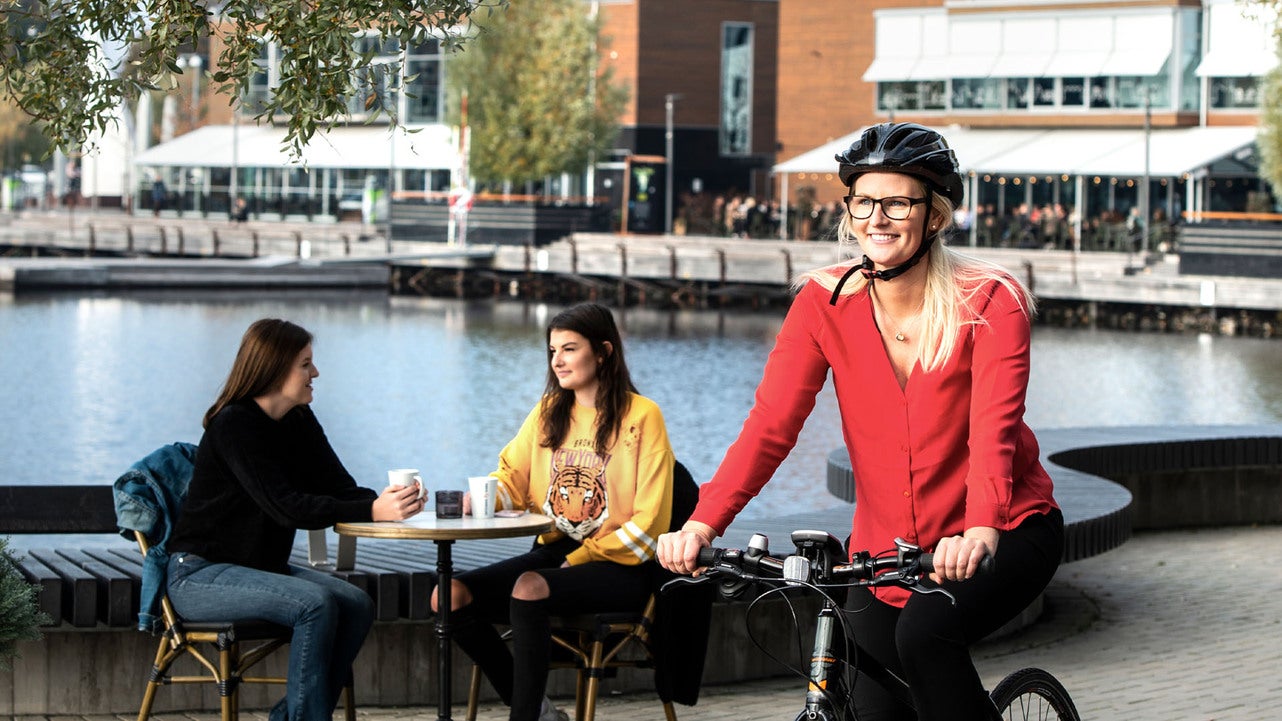 En kvinna med cykelhjälm och röd jacka cyklar förbi två unga kvinnor som sitter vid ett cafébord nära vattnet i centrala Jönköping. Höstträd och moderna byggnader syns i bakgrunden.