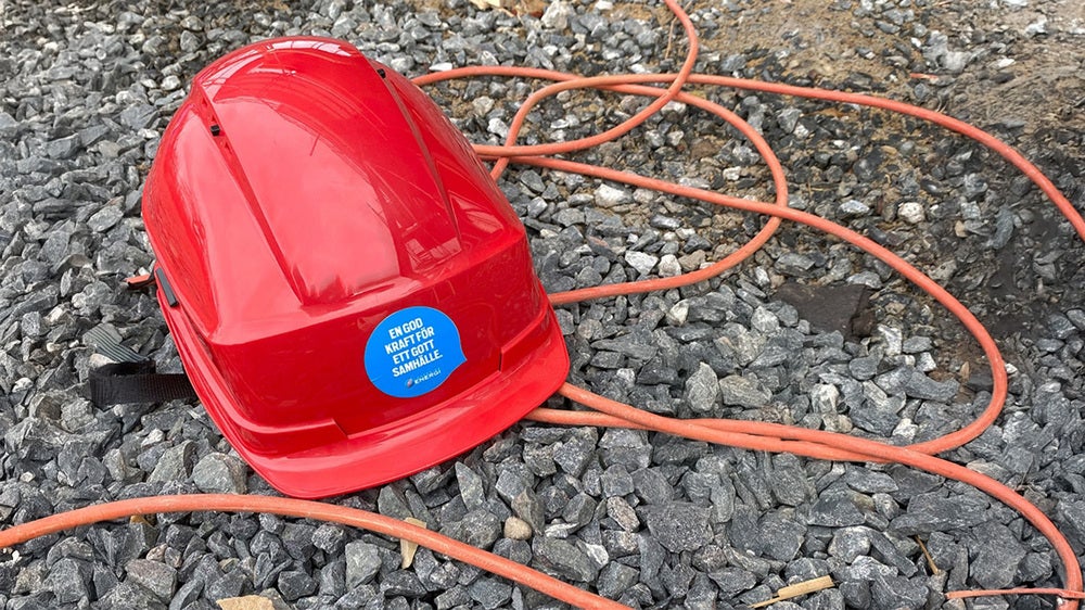 Röd skyddshjälm med blå klistermärke som ligger på grus tillsammans med orange elkablar.