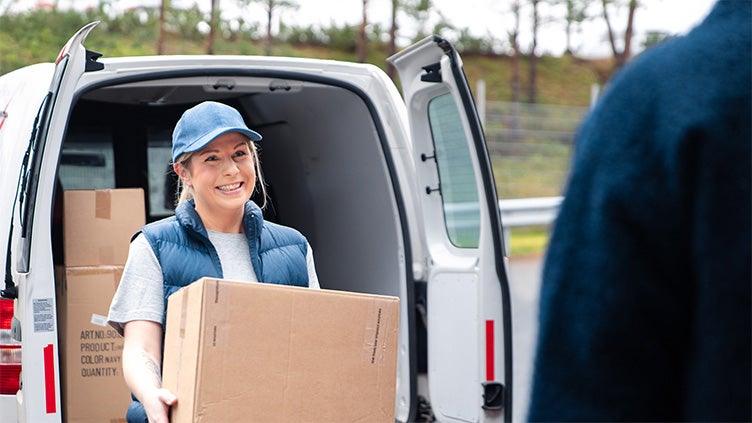 Person i keps och väst lastar ur paket från en vit transportbil vid en leverans, med fokus på mötet mellan leverantör och mottagare