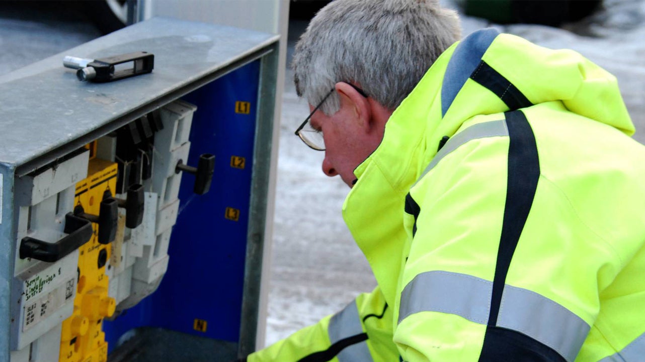 En elektriker i gul varseljacka arbetar med ett öppet elskåp. Skåpet innehåller flera strömbrytare och kopplingsenheter. Miljön är utomhus under kalla förhållanden, med frost på marken.