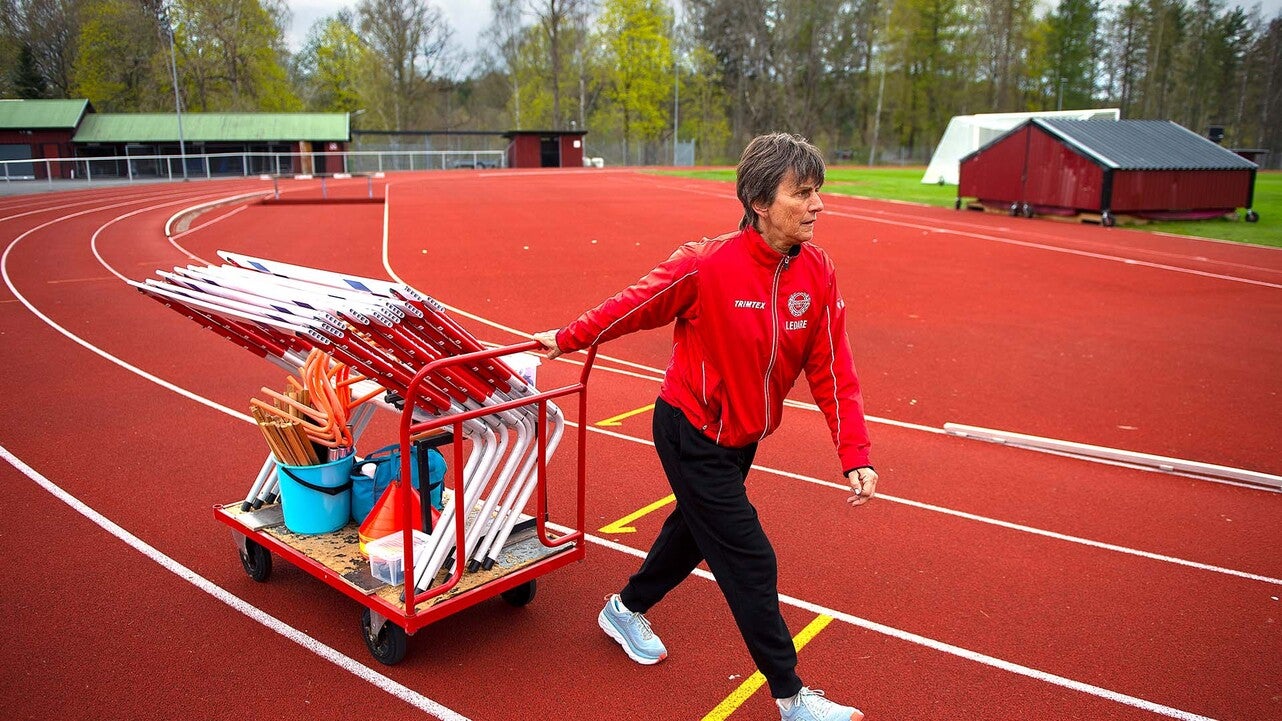 En person i röd tränarjacka drar en vagn fylld med idrottsutrustning över en röd löparbana, omgiven av grönskande träd och sportanläggningar i bakgrunden.