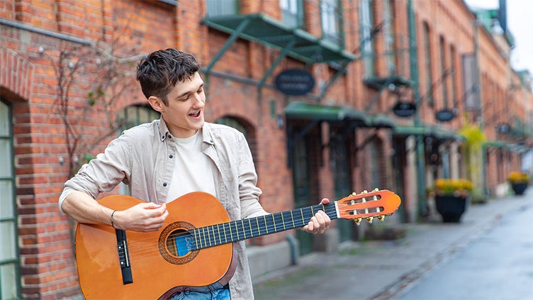 En ung man spelar akustisk gitarr på en gata kantad av röda tegelhus på Tändsticksområdet i Jönköping. 