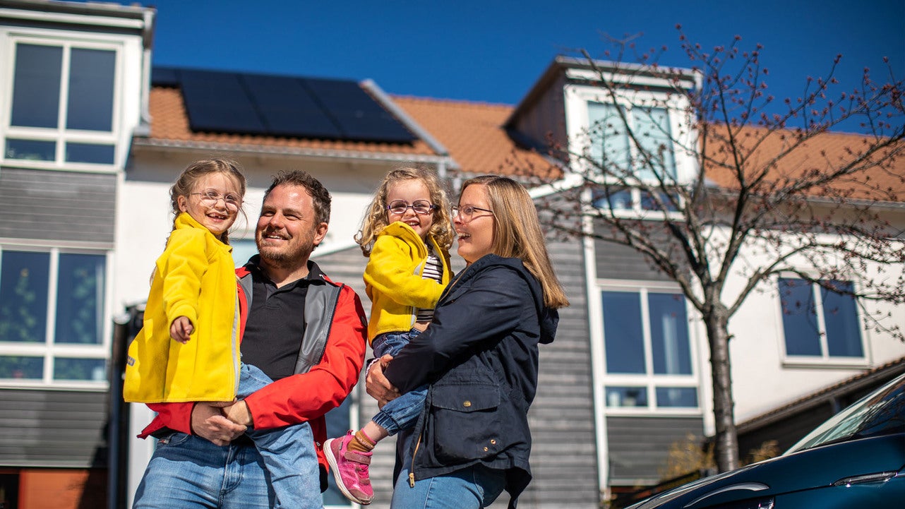 En glad familj med två föräldrar och två barn står framför sitt moderna hus med solpaneler på taket. Solen skiner och familjen är klädd i färgglada jackor. Huset har stora fönster och röda takpannor.