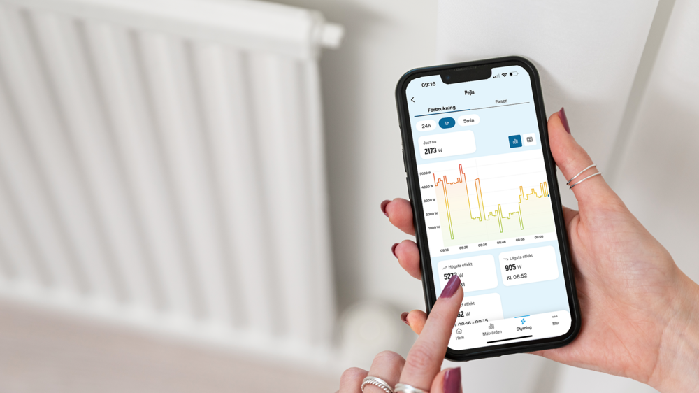Person använder Jönköping Energis app för att följa elförbrukning i realtid via färgkodad linjegraf, med ett element synligt i bakgrunden.