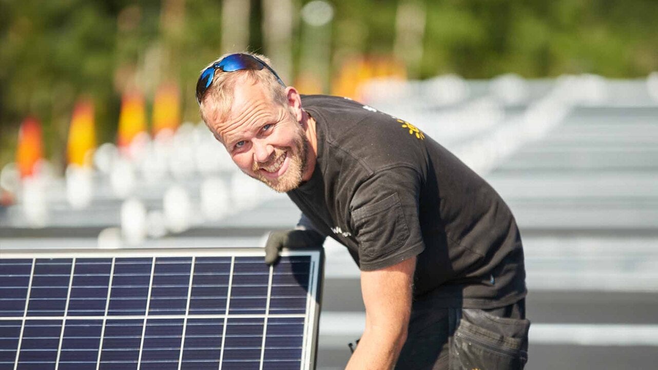 En leende installatör arbetar med att justera en solpanel på en solenergianläggning utomhus i solsken.