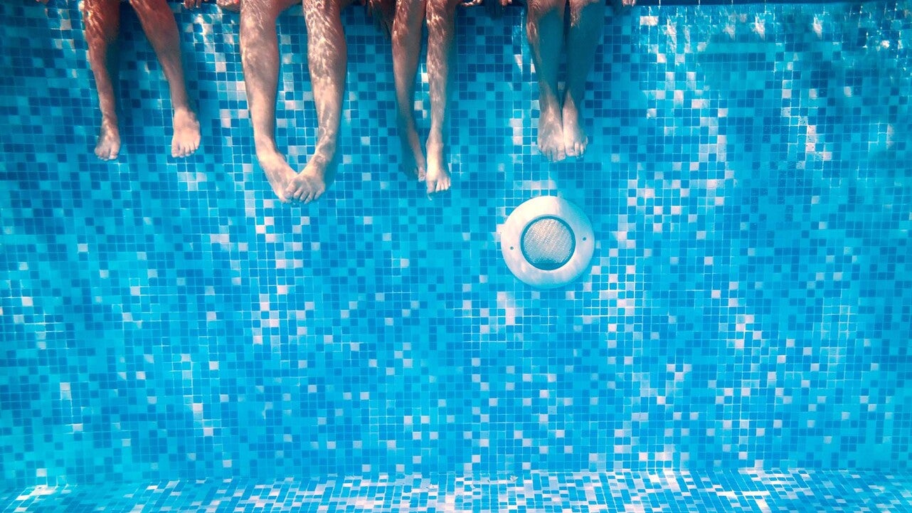 Flera personers ben som dinglar över kanten av en pool, med en klarblå mosaikvägg och vatten som reflekterar solljus. En rund poollampa syns i väggen under vattnet.