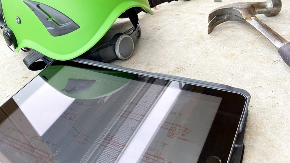 En surfplatta som visar byggnadsritningar ligger på marken bredvid en grön hjälm och en hammare. Bilden föreslår en byggarbetsmiljö där digital teknik används.