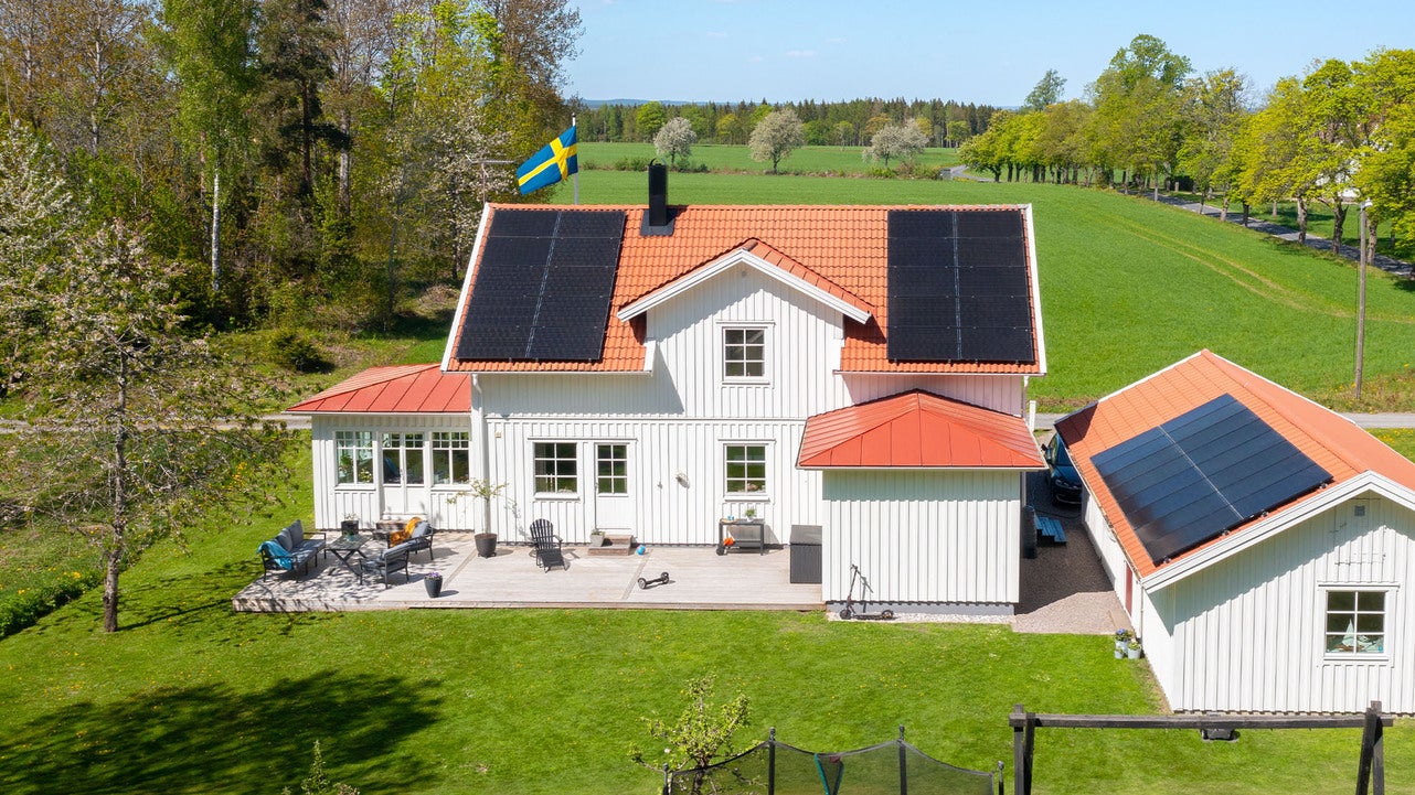 Ett vitt hus på landsbygden med solpaneler installerade på taket både på huvudbyggnaden och garaget. En svensk flagga vajar på taket, och huset är omgivet av grönska och öppna fält.