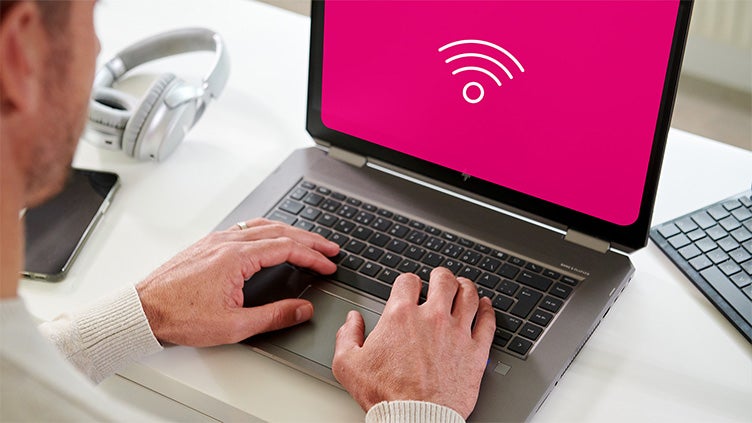 En person sitter vid en laptop med en rosa skärm som visar en vit Wi-Fi-symbol, vilket indikerar internetuppkoppling. Bredvid datorn ligger hörlurar på skrivbordet.
