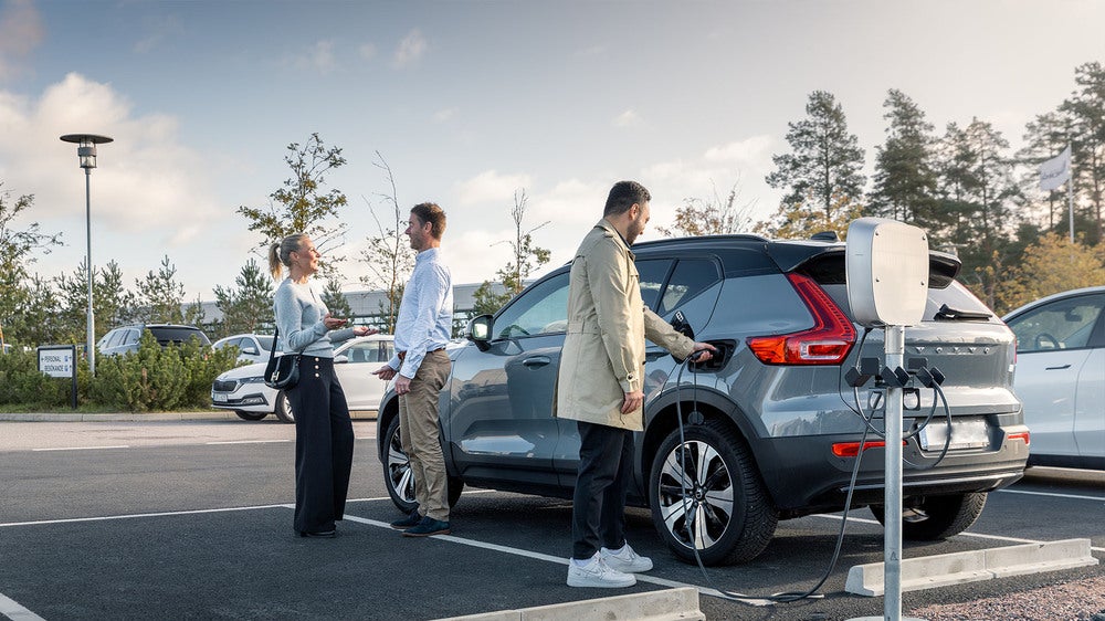 Tre personer står vid en grå SUV som laddas vid en elbilsladdstation på en parkeringsplats. En man ansluter laddkabeln till bilen medan de andra två samtalar bredvid. I bakgrunden syns fler parkerade bilar och träd under en molnig himmel.