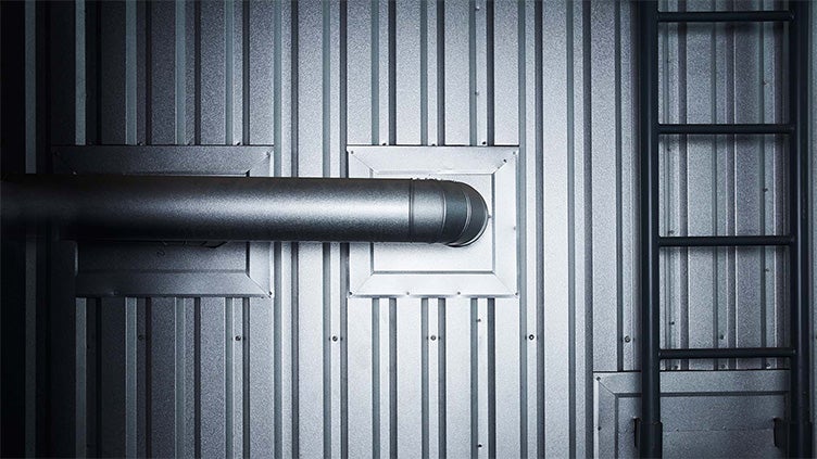Ett rör passerar genom en metallklädd industrivägg via en fyrkantig genomföring. Till höger syns en vertikal stege.