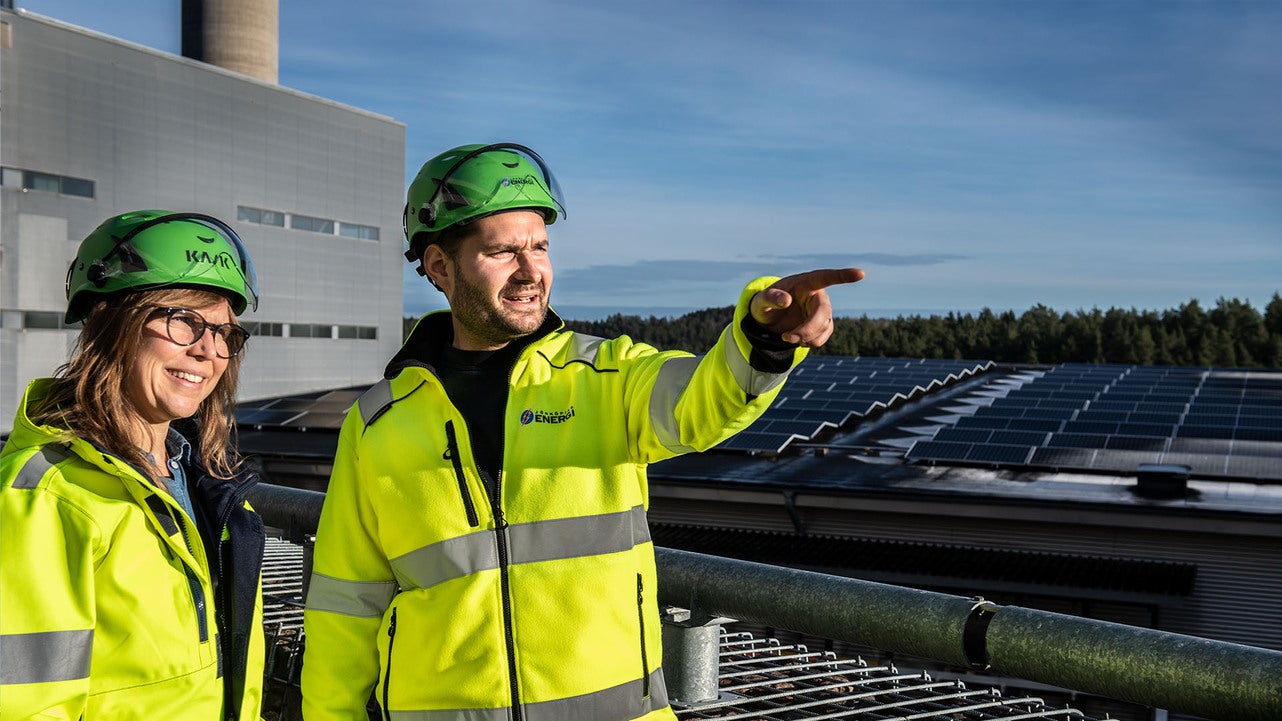 Två tekniker från Jönköping Energi, klädda i gula varseljackor och gröna hjälmar, står vid en solcellsanläggning. Den ena teknikern pekar mot något i fjärran medan de diskuterar arbetet. Solpaneler och en industribyggnad syns i bakgrunden.