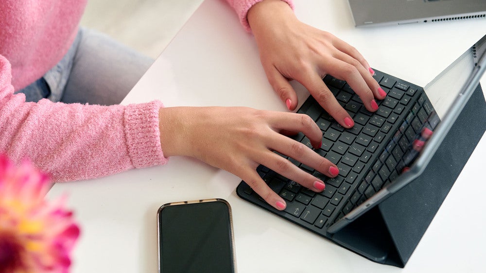 En person i rosa tröja skriver på ett tangentbord anslutet till en surfplatta, medan en smartphone ligger bredvid på skrivbordet. En blomma syns i förgrunden.