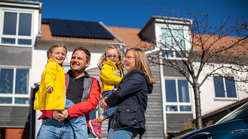 Leende familj med två barn står utanför sitt radhus, där solpaneler är installerade på taket under en klarblå himmel.