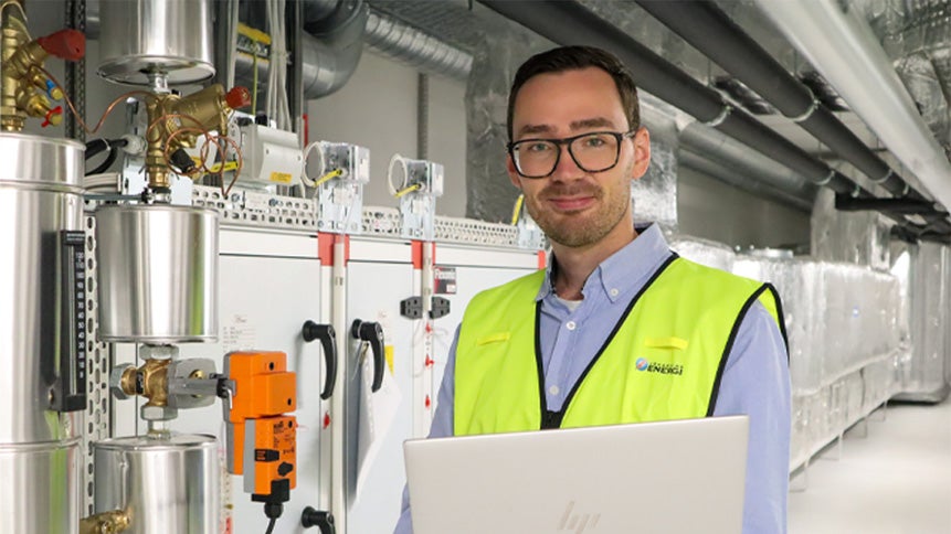 En manlig energikonsult i ljusblå skjorta och varselväst står med en laptop i famnen och ler mot kameran omgiven av rör, skåp och kablar i en teknisk miljö.