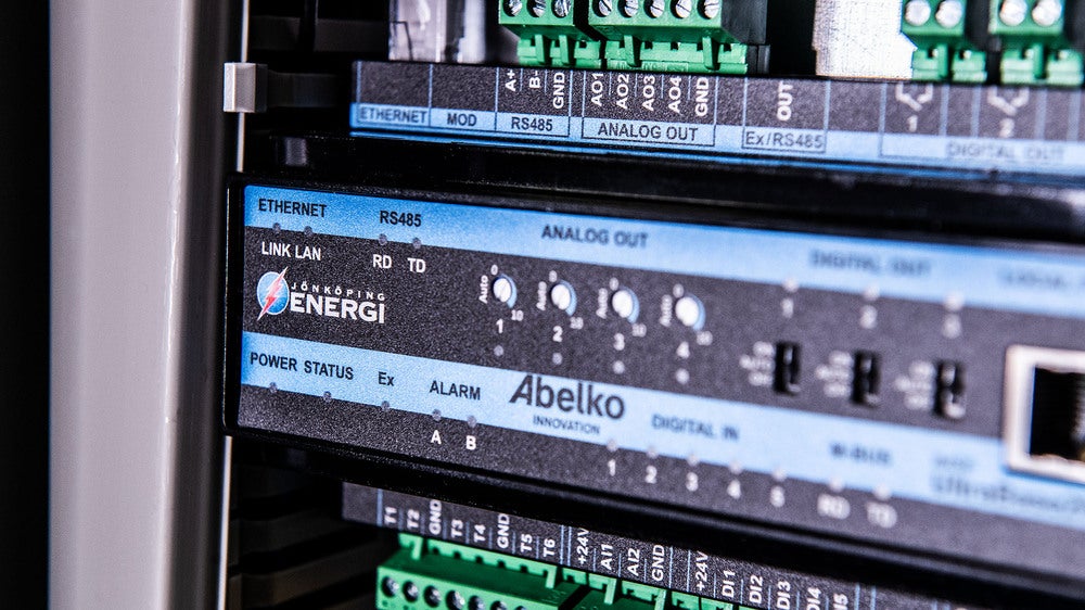 En närbild på nätverks- och kontrollutrustning som används av Jönköping Energi. Panelen har flera anslutningar för Ethernet, RS485 och analoga utgångar, med statuslampor och märket "Abelko" tydligt synligt.