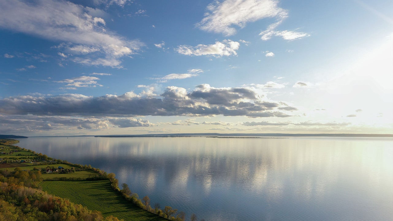 En storslagen vy över sjön Vättern under en klar himmel med moln, där vattnet speglar himlen. Solen lyser mjukt över landskapet, och i förgrunden syns grönskande mark längs sjöns kant.