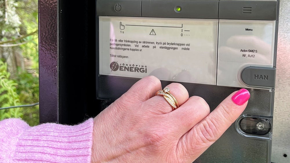 Hand med rosa nagellack trycker på HAN-knappen på en digital elmätare märkt Jönköping Energi, med display och informationstext synlig.