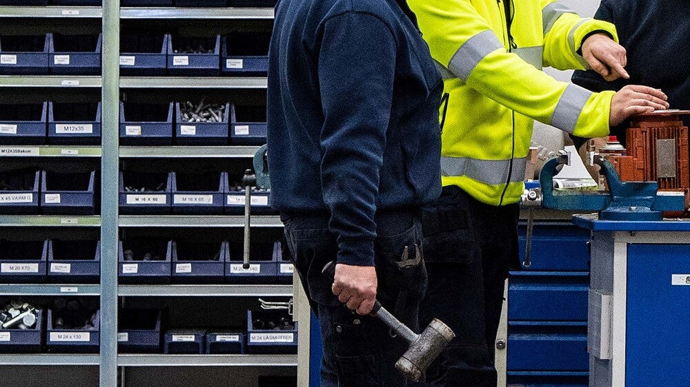 Två industrimedarbetare, en i mörkblå arbetskläder och en i gul reflexjacka, arbetar i en verkstad. En av dem håller en gummiklubba, medan hyllor fyllda med sorterade skruvar och komponenter syns i bakgrunden.