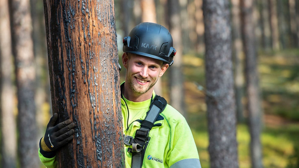 En leende tekniker iförd hjälm och arbetskläder står lutad mot en elstolpe i en skog, med solljus som lyser upp den omgivande naturen.