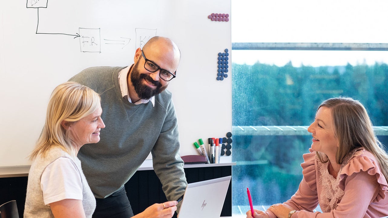 Tre medarbetare från Jönköping Energi deltar i ett möte. De sitter vid ett bord med en bärbar dator och diskuterar, medan en whiteboard med ritade diagram syns i bakgrunden. Genom fönstren syns en vy över grönskande träd.