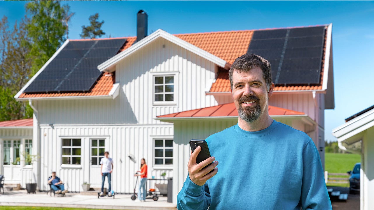 En man i blå tröja står leende utomhus och håller en smartphone framför ett vitt hus med solpaneler på taket. I bakgrunden syns barn som leker med en sparkcykel och en cykel.