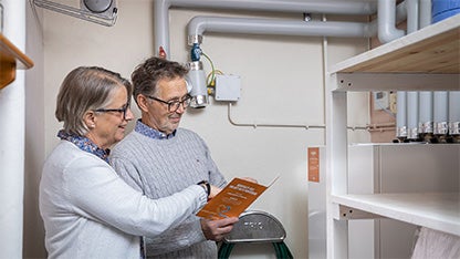 Äldre par står i ett bostadsutrymme med synlig rördragning och fjärrvärmeutrustning
