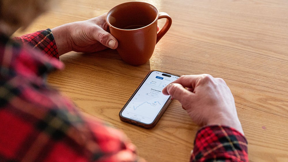 En person i rutig skjorta håller i en kopp kaffe och använder en smartphone för att övervaka energiförbrukning via en mobilapp. Skärmen visar en graf över förbrukningsdata.