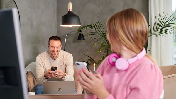En pappa i ljus tröja sitter i soffan med sin mobil i handen och en laptop framför sig, medan han tittar på sin dotter. Dottern, klädd i en rosa tröja och hörlurar, håller en mobiltelefon och har en skärm framför sig. Båda är uppkopplade och surfar via fibernät i vardagsmiljö.