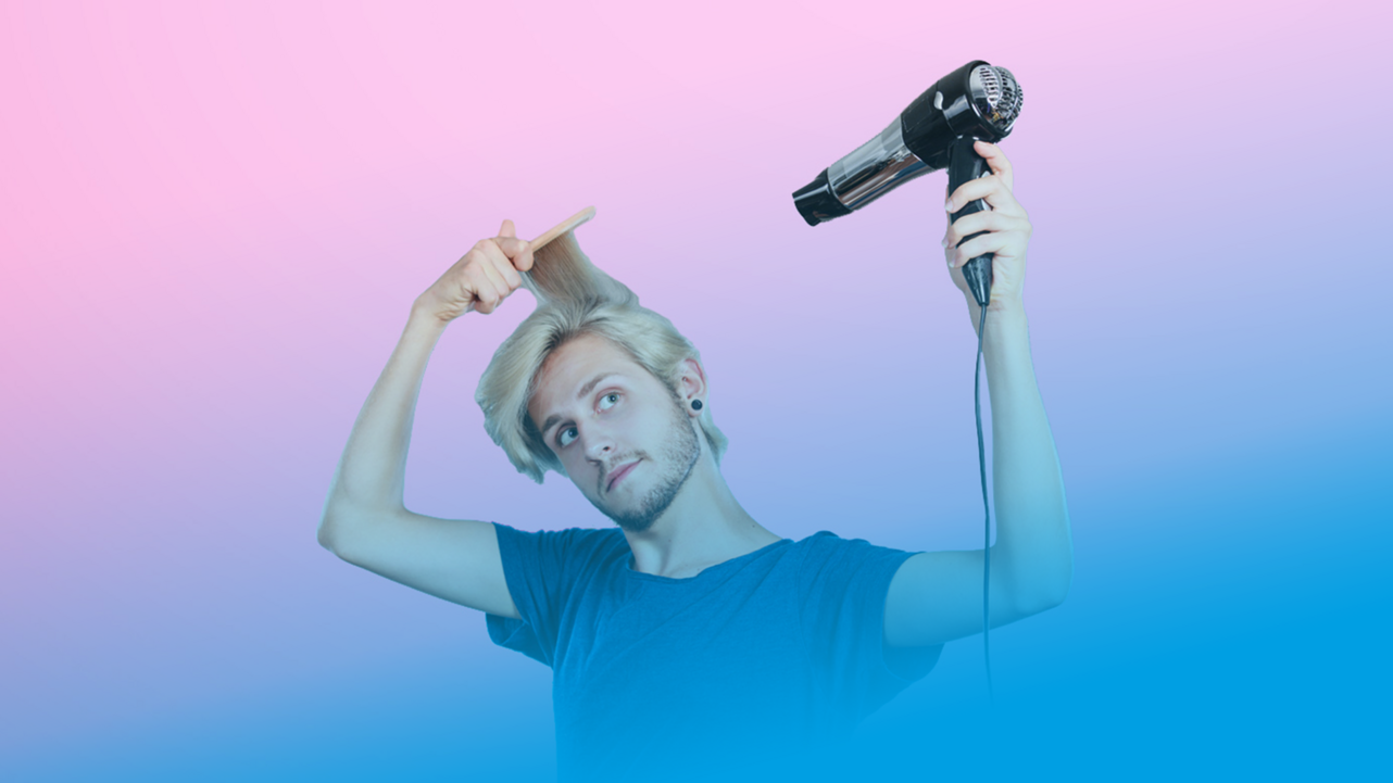 En man med blont hår och blå t-shirt använder en hårfön och kam för att styla sitt hår. Han står framför en rosa bakgrund.