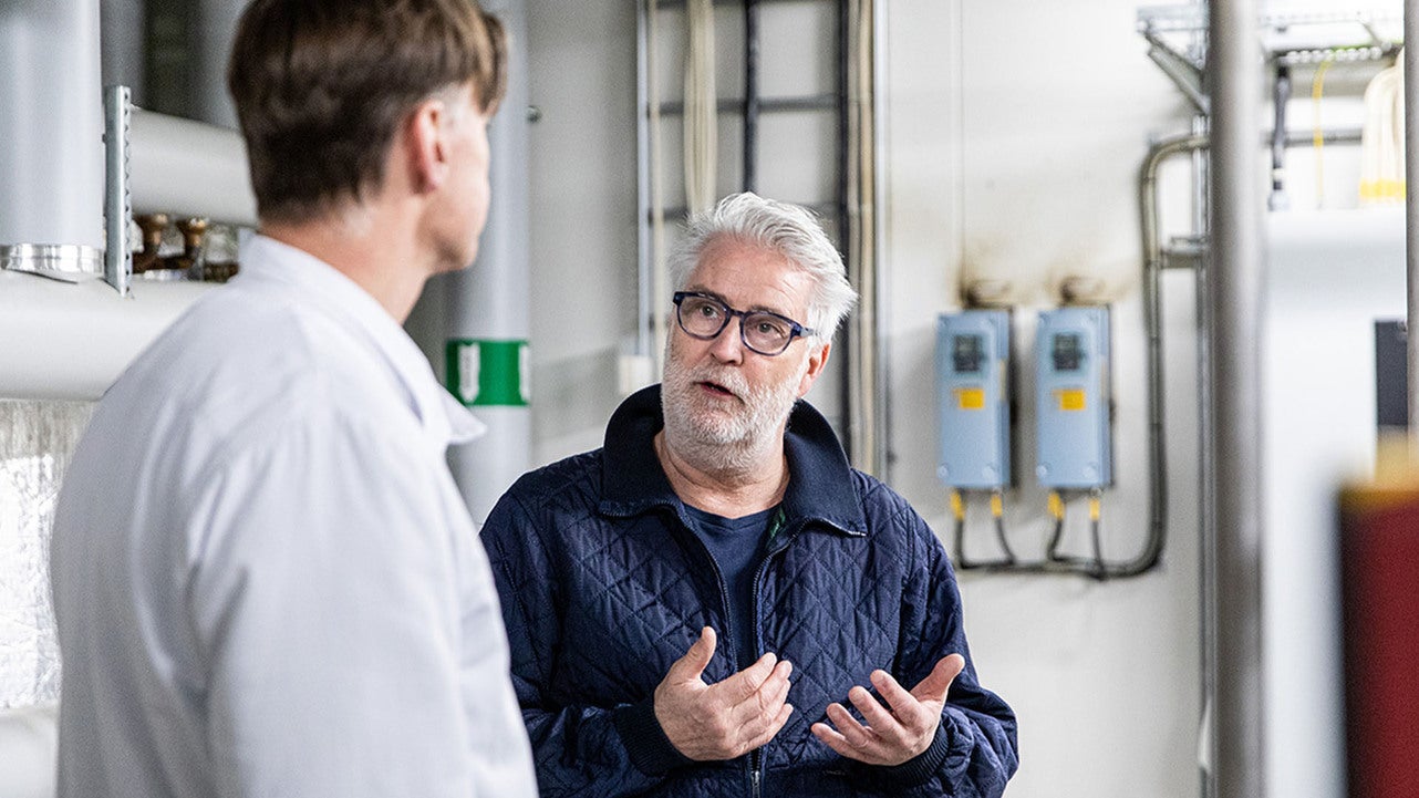 En man i blå arbetsjacka gestikulerar under en konversation med en man i vit labbrock, medan de står bland maskiner inne på Danish Crowns anläggning.