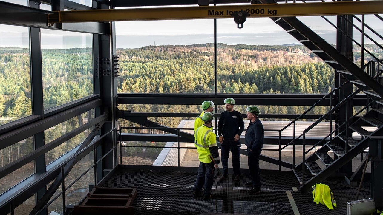 Tre tekniker i skyddsutrustning står och samtalar på en hög nivå i en industriell energianläggning på kraftvärmeverket på Torsvik med stora fönster som visar utsikt över ett skogsområde. Metalltrappor och en lyftanordning syns i förgrunden.