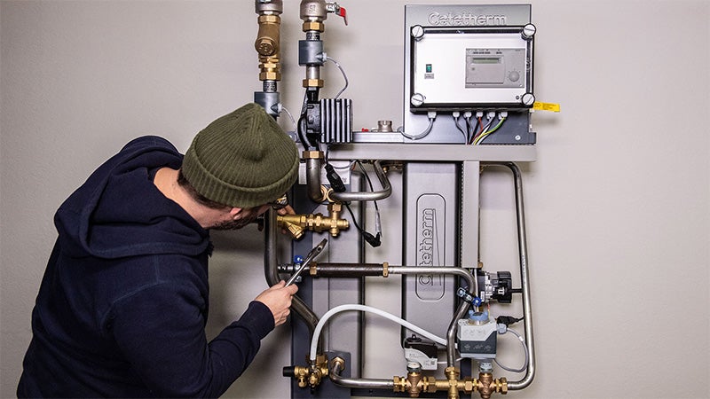 Person i mössa arbetar med verktyg vid en väggmonterad fjärrvärmecentral med rörsystem, ventiler, mätutrustning och styrbox.