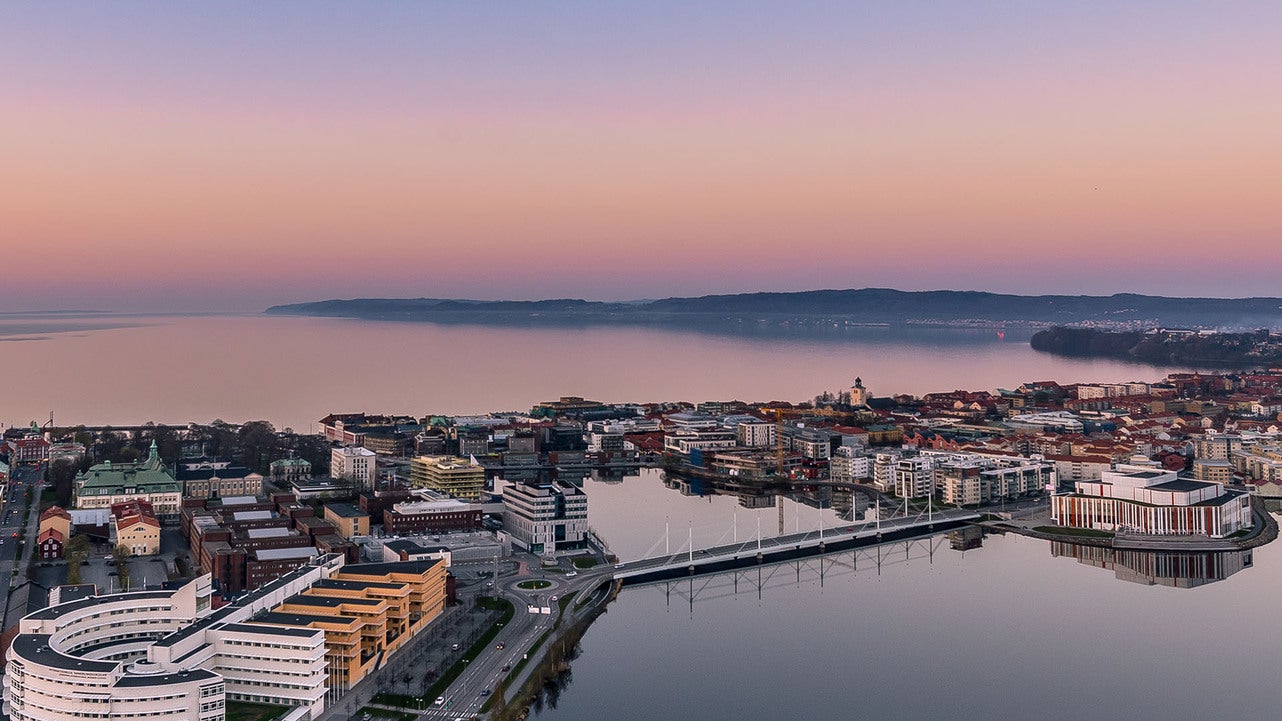 Flygfoto över centrala Jönköping vid Vätterns strand under solnedgången. Bilden visar stadens moderna byggnader, universitet, bron över Munksjön och det stilla vattnet med en färggrann himmel i bakgrunden.