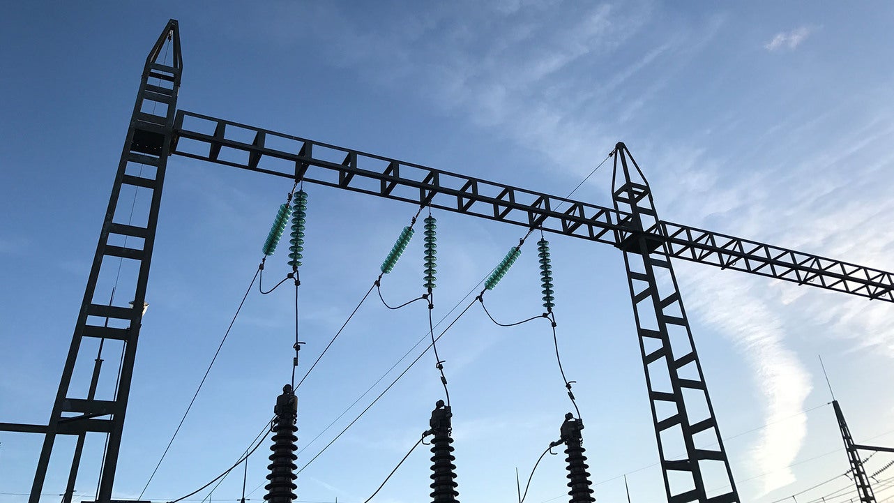 Elstation med flera kraftledningsstolpar och gröna isolatorer upphängda på en metallställning under en klar himmel. Ledningarna är spända mellan stolparna, som del av en högspänningsanläggning.