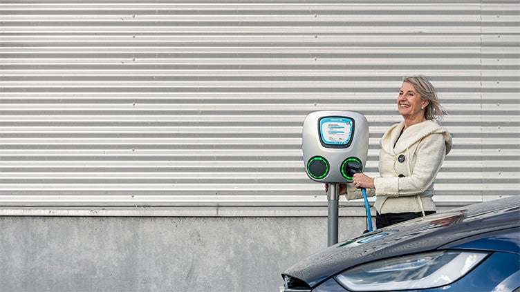 En kvinna i ljus jacka laddar sin elbil vid en laddstation utomhus. Hon håller i laddkabeln och ser leende ut medan hon står bredvid bilen. Bakgrunden består av en vägg med metallpaneler.