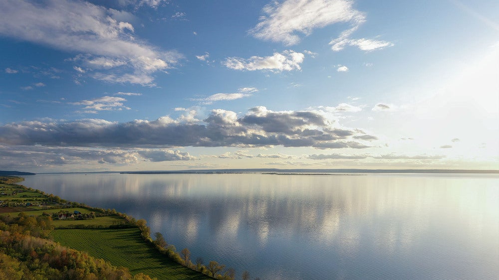 En storslagen vy över sjön Vättern under en klar himmel med moln, där vattnet speglar himlen. Solen lyser mjukt över landskapet, och i förgrunden syns grönskande mark längs sjöns kant.