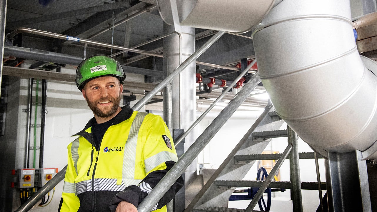 En installatör med grön arbetshjälm och varselkläder går nedför en metalltrappa i en teknisk anläggning. Han är omgiven av ett komplext nätverk av rör och elektriska knappboxar som styr fjärrkylanläggningen.