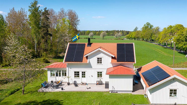 Vit villa med rött tegeltak och installerade solpaneler på både boningshus och garage, omgiven av grönskande fält och svensk flagga på taket.