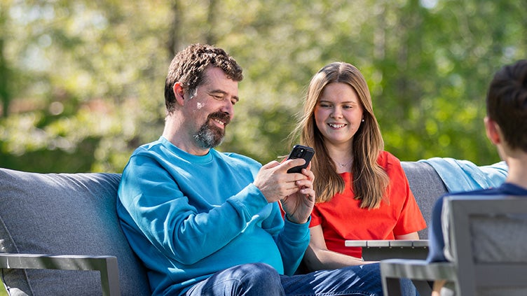 Man i blå tröja visar en kvinna i röd tröja något i mobilen. De sitter på en altan med grön skog bakom.