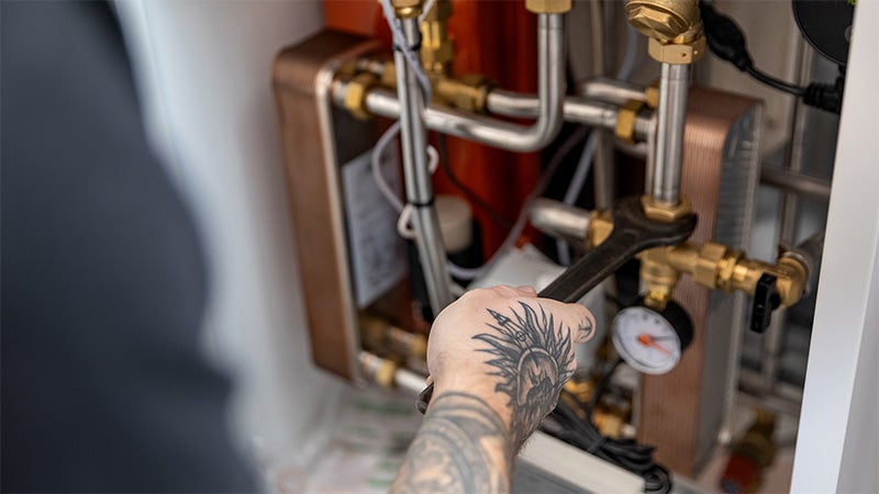 Person med tatuering använder rörtång för att justera koppling på fjärrvärmecentral med kopparrör, värmeväxlare och tryckmätare.