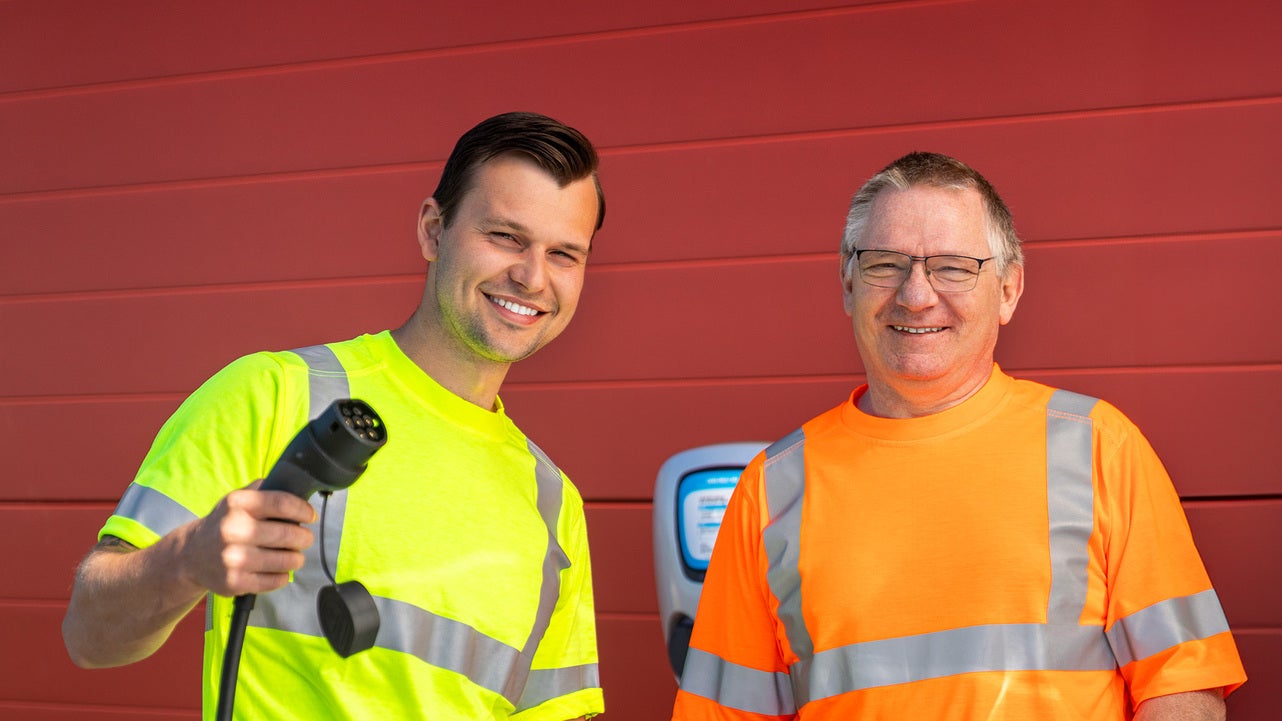 Två tekniker från Jönköping Energi, klädda i neongula och orange arbetskläder, står framför en elbilsladdningsstation. Den ena teknikern håller i en laddkabel och båda ler mot kameran, med en röd byggnadsvägg i bakgrunden.