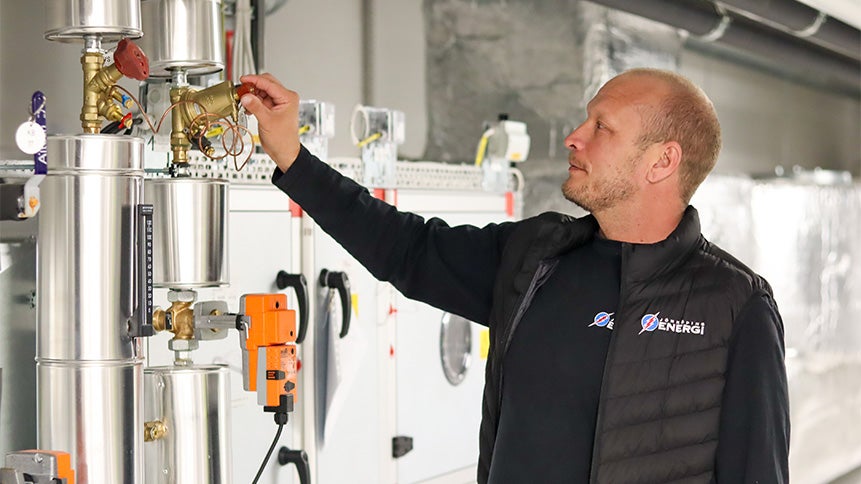 En man står vid en mätare och inspekterar en industriell, teknisk miljö med stora rör och teknisk utrustning.